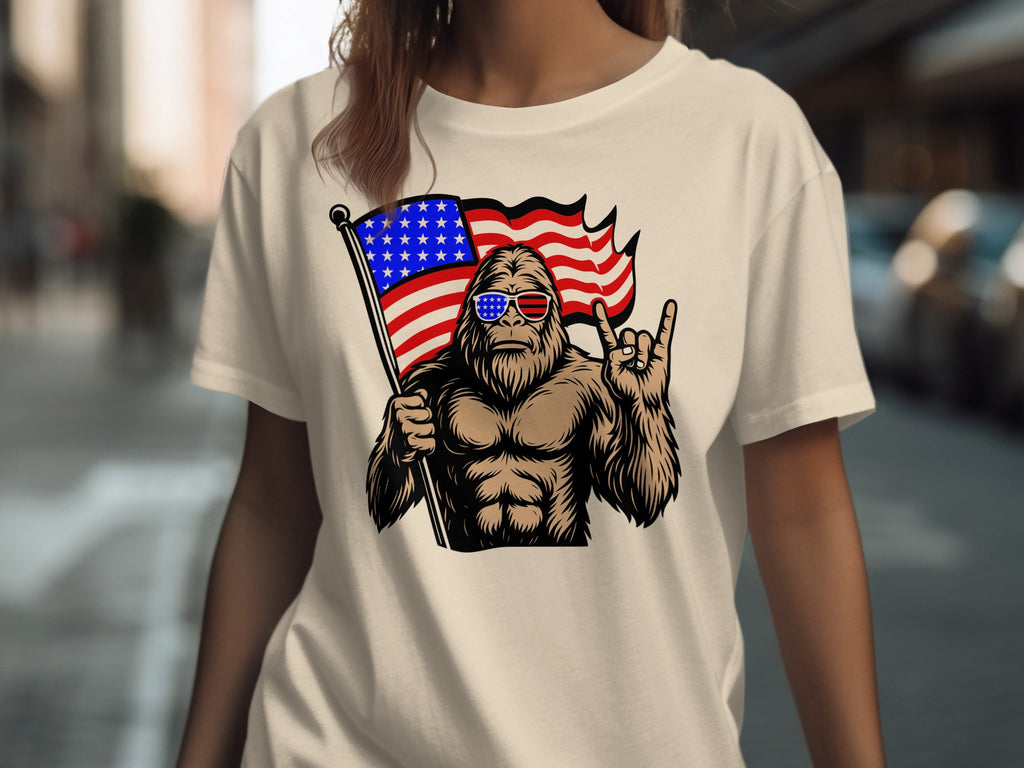 Bigfoot with American flag sunglasses holding US flag, making rock on hand gesture.