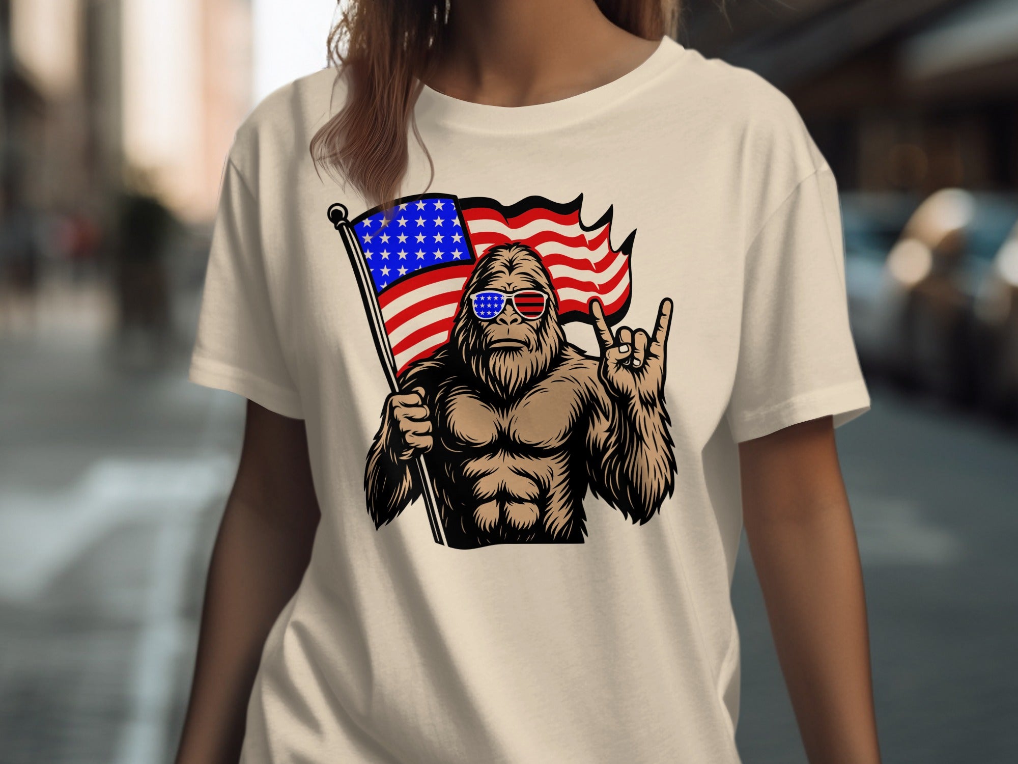 Bigfoot with American flag sunglasses holding US flag, making rock on hand gesture.