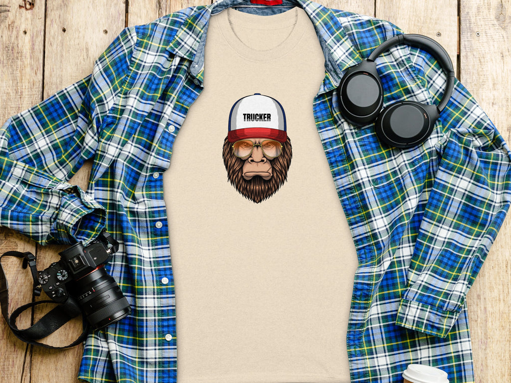 Beige t-shirt graphic of a Bigfoot wearing a "Trucker" hat and aviator glasses.