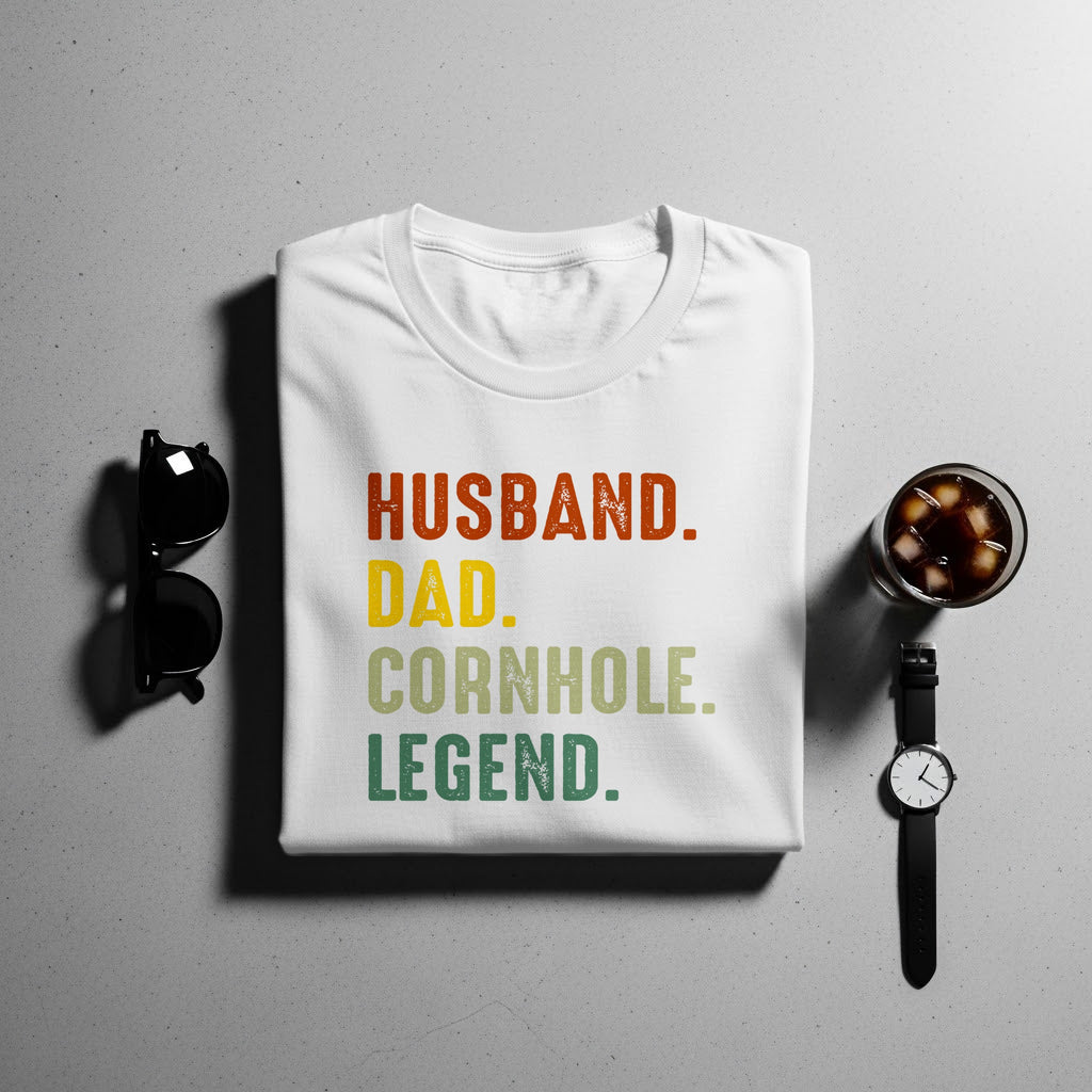 White t-shirt with "HUSBAND. DAD. CORNHOLE. LEGEND." text in gradient colors.