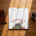 White t-shirt with graphic of a frog reading under blankets, text "ONE CHAPTER IN...".
