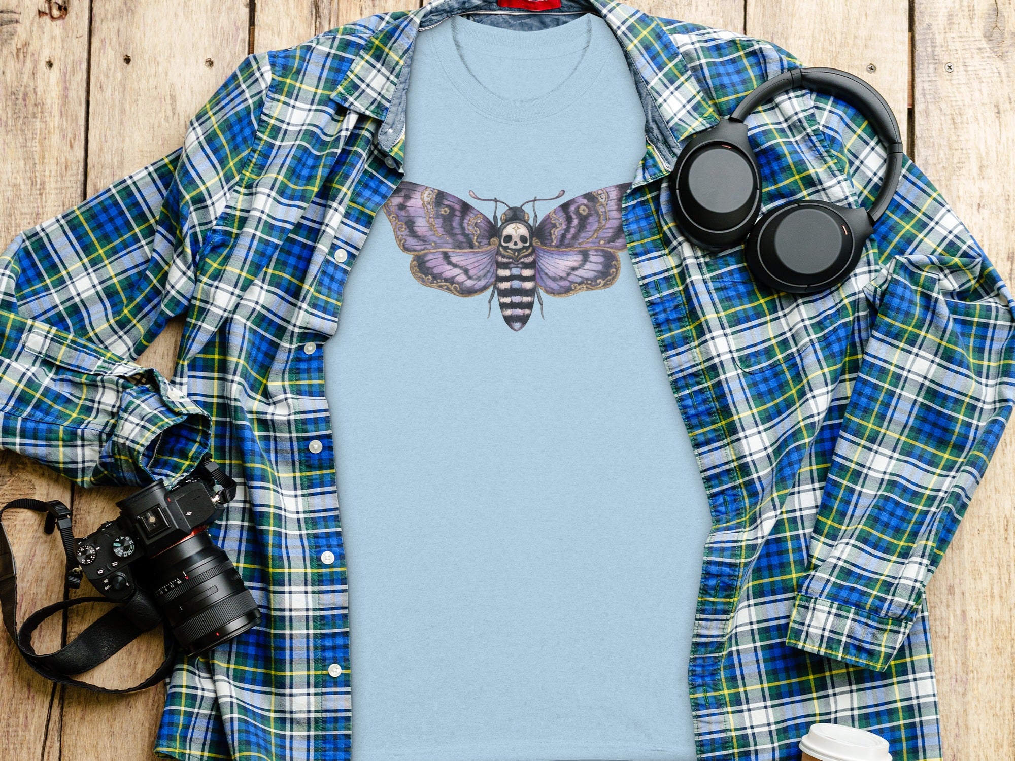 Light blue t-shirt with purple moth skull graphic, layered under a blue plaid shirt.