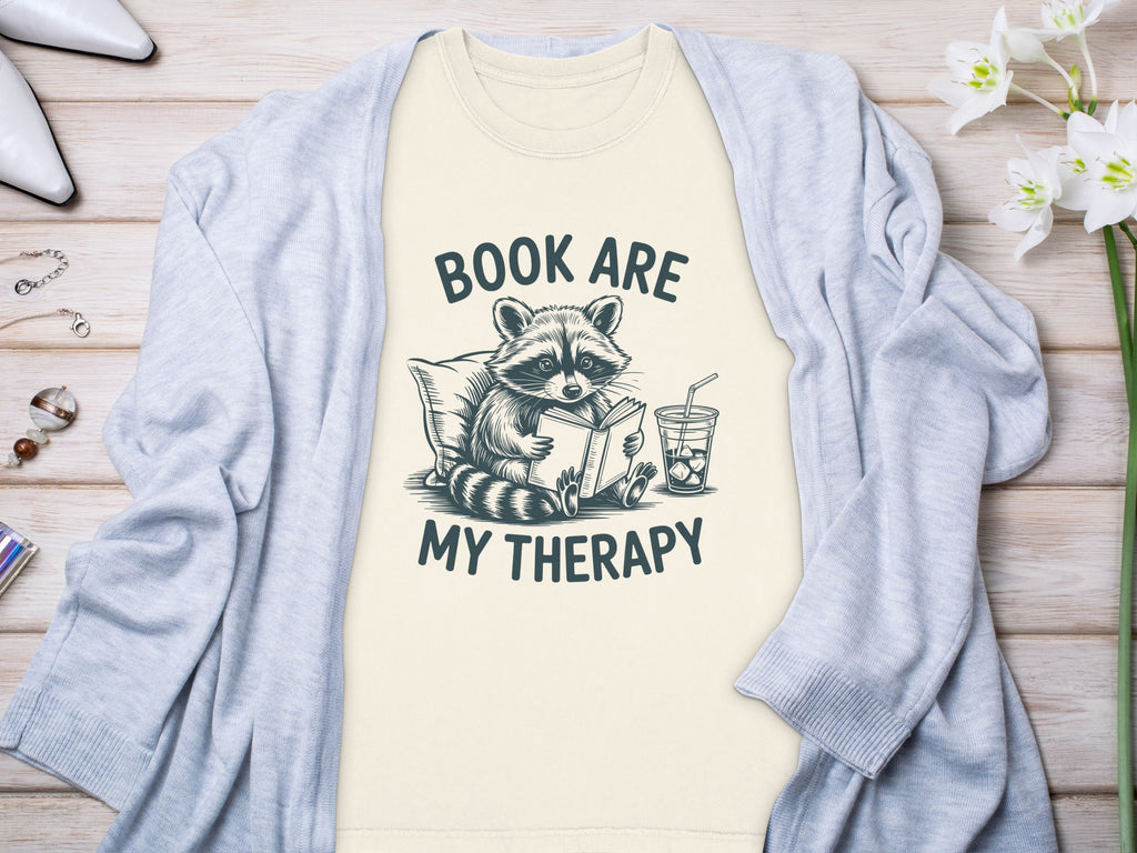 T-shirt with raccoon reading, "Book Are My Therapy" text, and iced drink.
