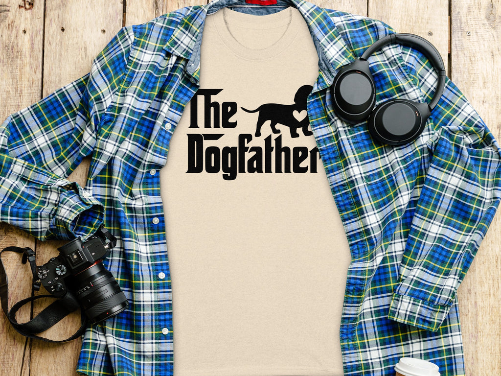 T-shirt with "The Dogfather" text and dog silhouette, laid flat with flannel shirt and camera.