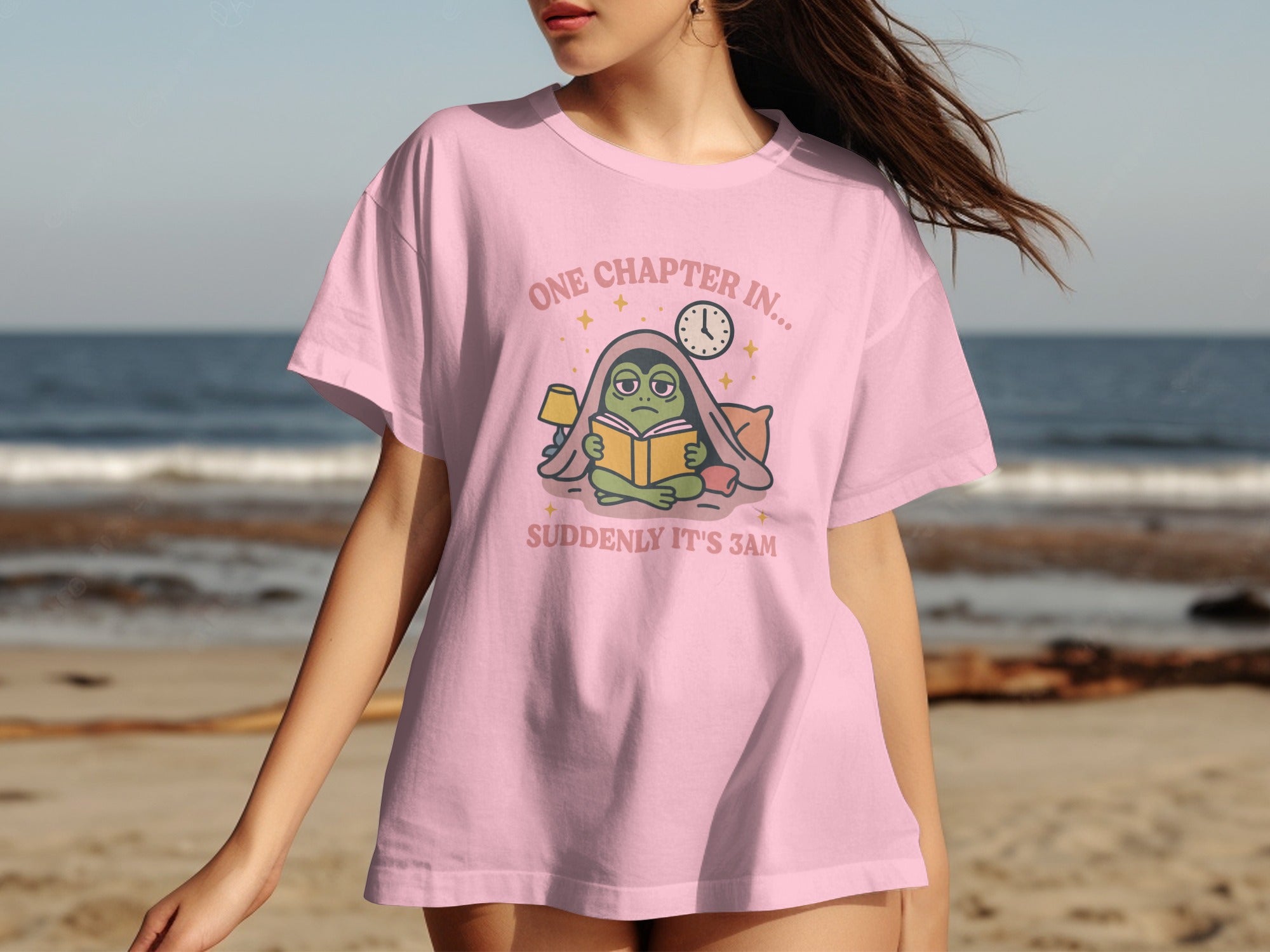 Pink t-shirt graphic featuring a tired frog reading a book, with text "ONE CHAPTER IN... SUDDENLY IT'S 3AM".
