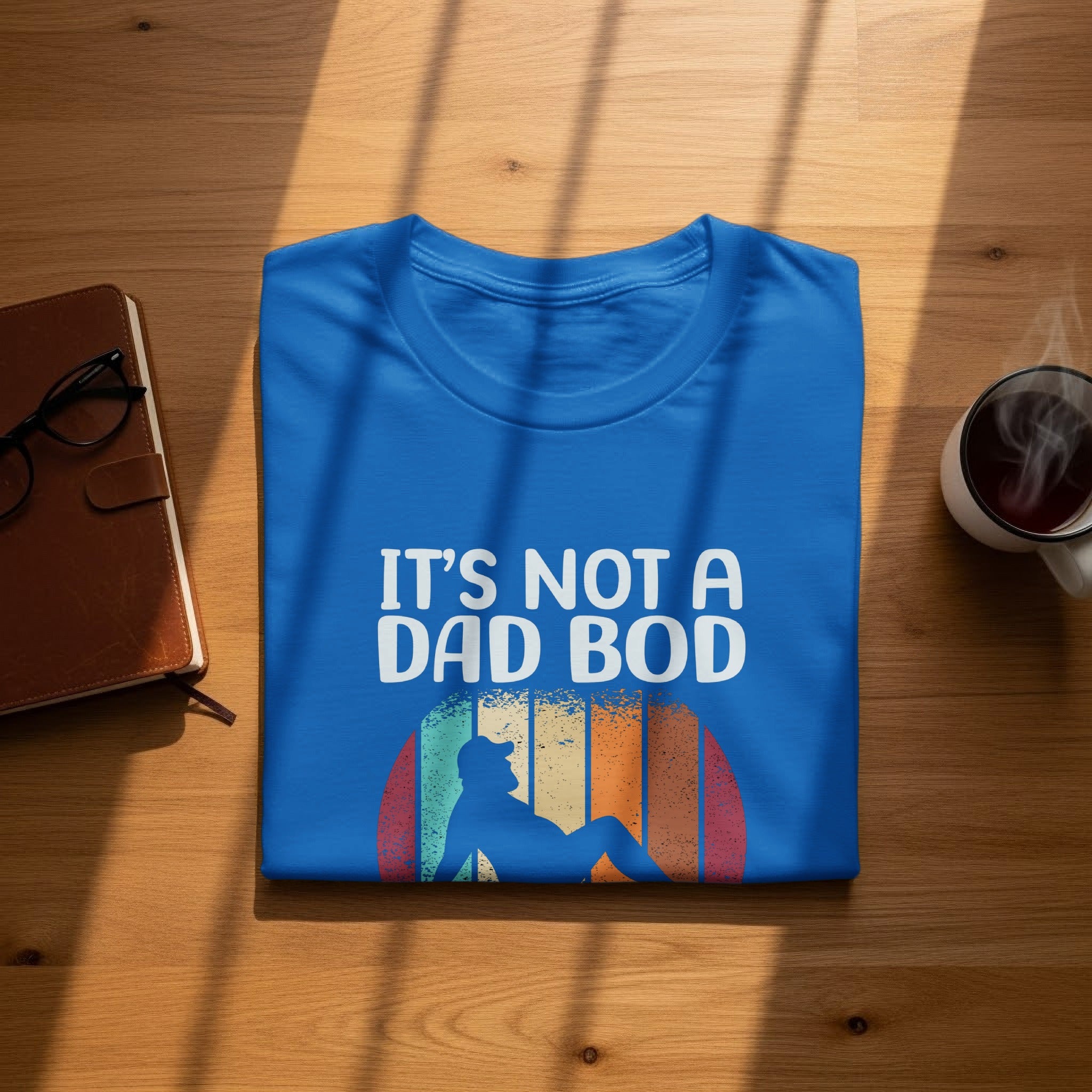 Blue t-shirt with "IT'S NOT A DAD BOD" text and silhouette against sunset stripes.