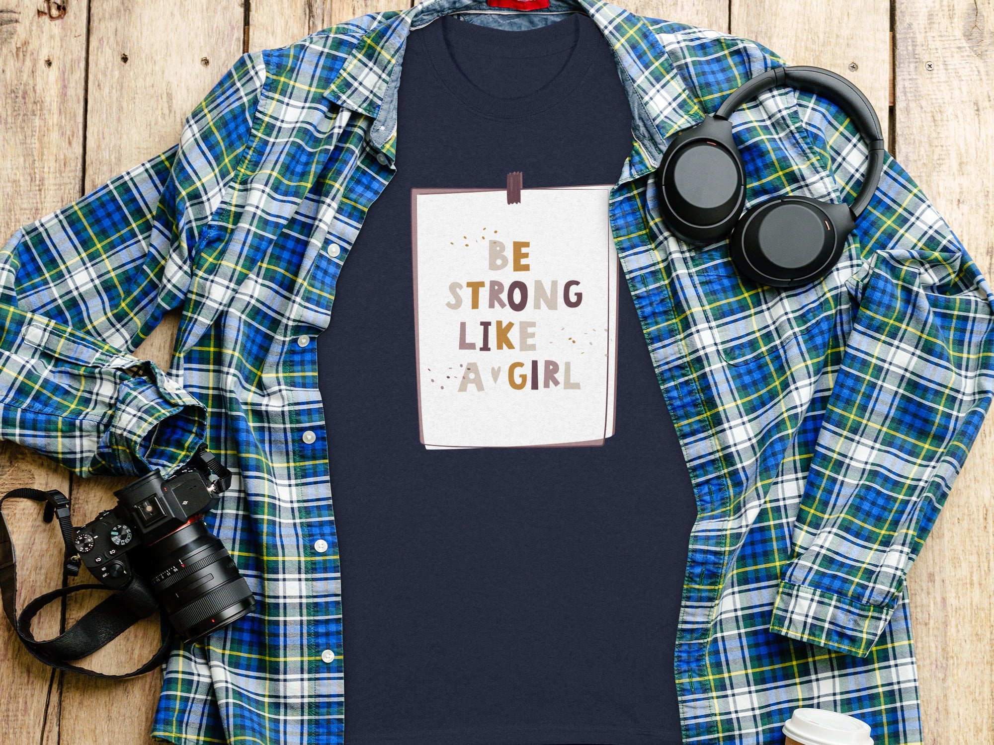 Navy t-shirt with text "BE STRONG LIKE A GIRL" laid flat with plaid shirt and headphones.