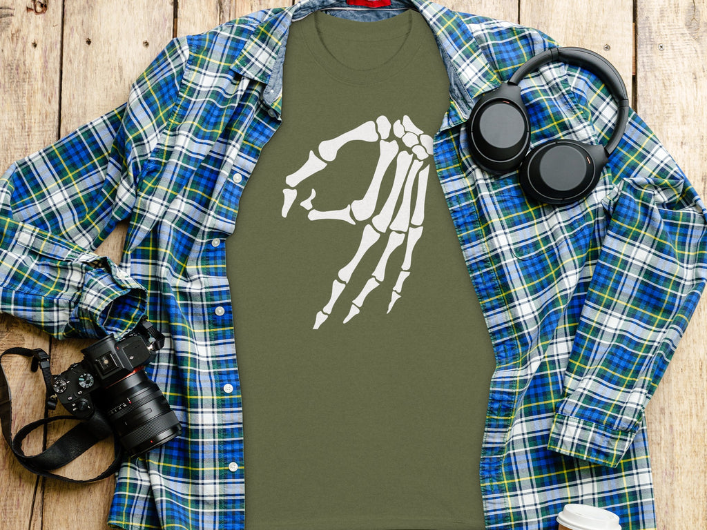 Olive green t-shirt with white skeleton hand graphic, plaid shirt, headphones, and camera.