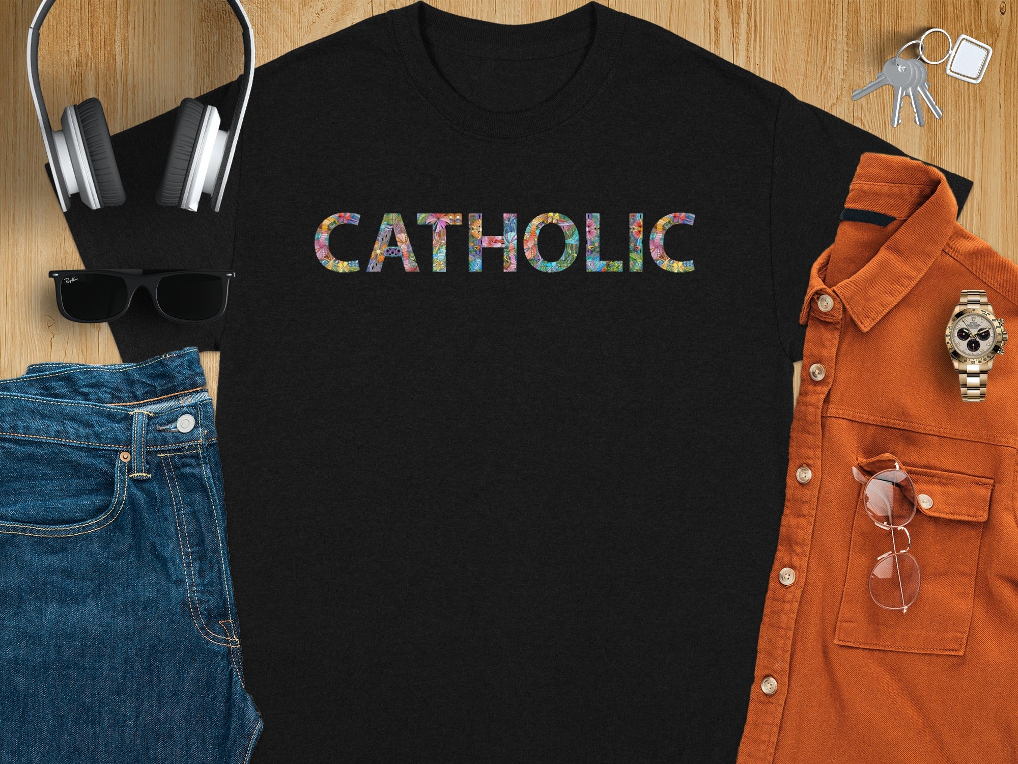 Black t-shirt with colorful "CATHOLIC" text, flat lay with headphones, jeans, and orange shirt.