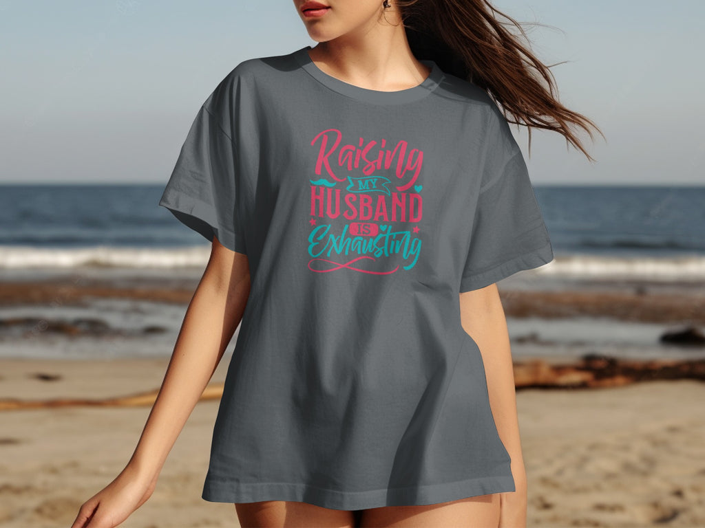 Raising my husband is exhausting text on a gray t-shirt on a beach.