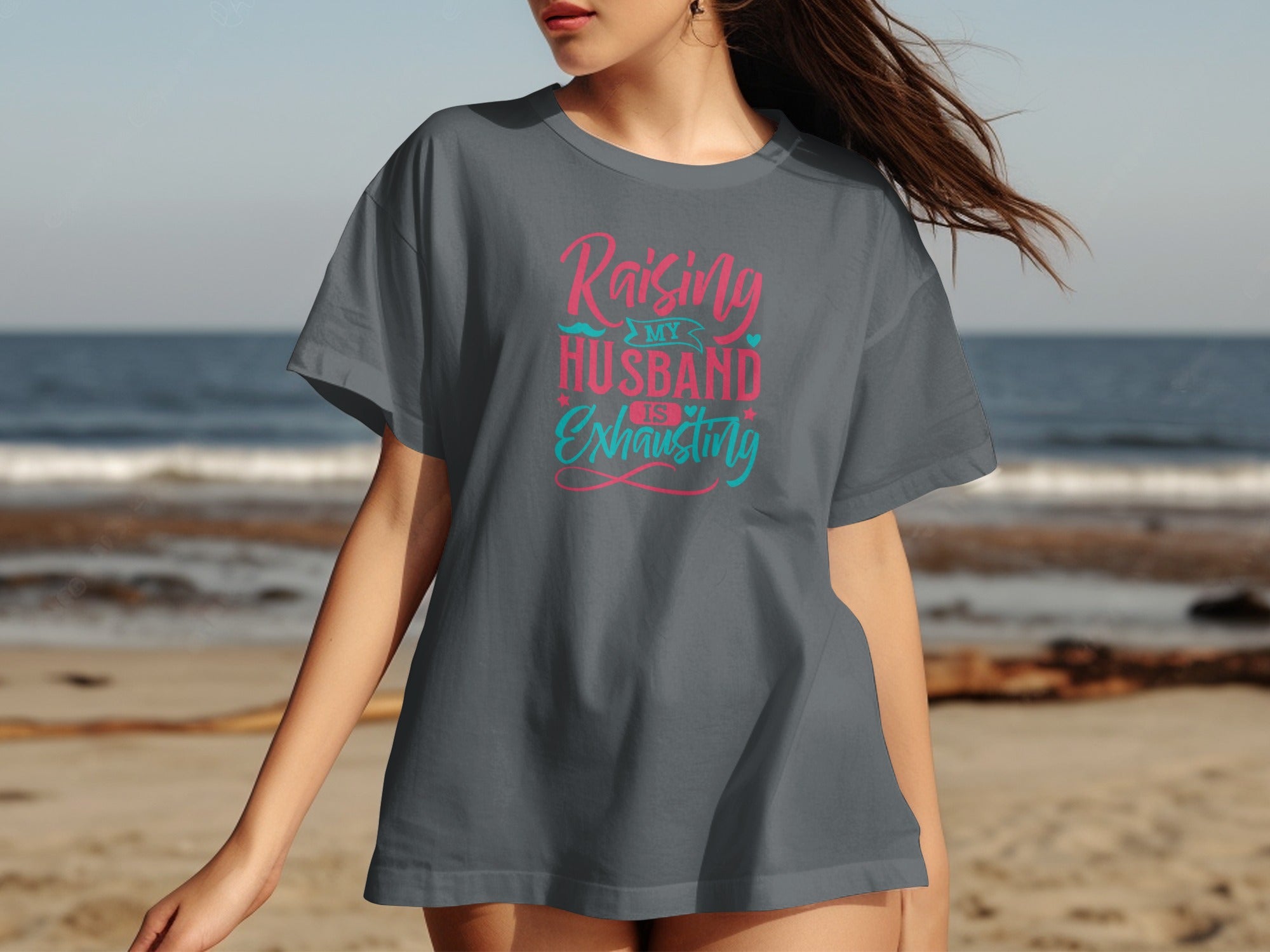 Raising my husband is exhausting text on a gray t-shirt on a beach.