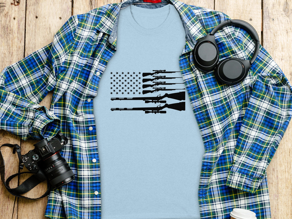 Light blue t-shirt with American flag made of rifles, laid on a wooden surface.