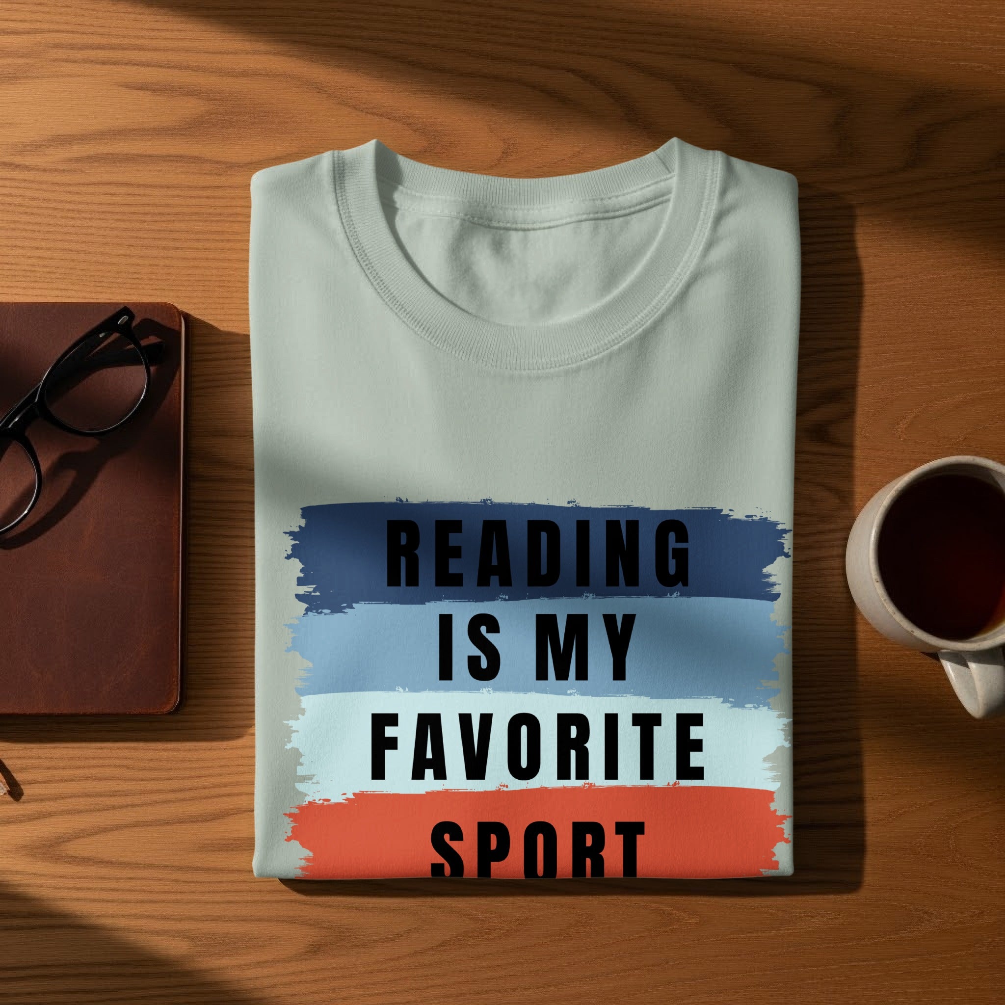 Light blue t-shirt with "READING IS MY FAVORITE SPORT" text on a wooden table.