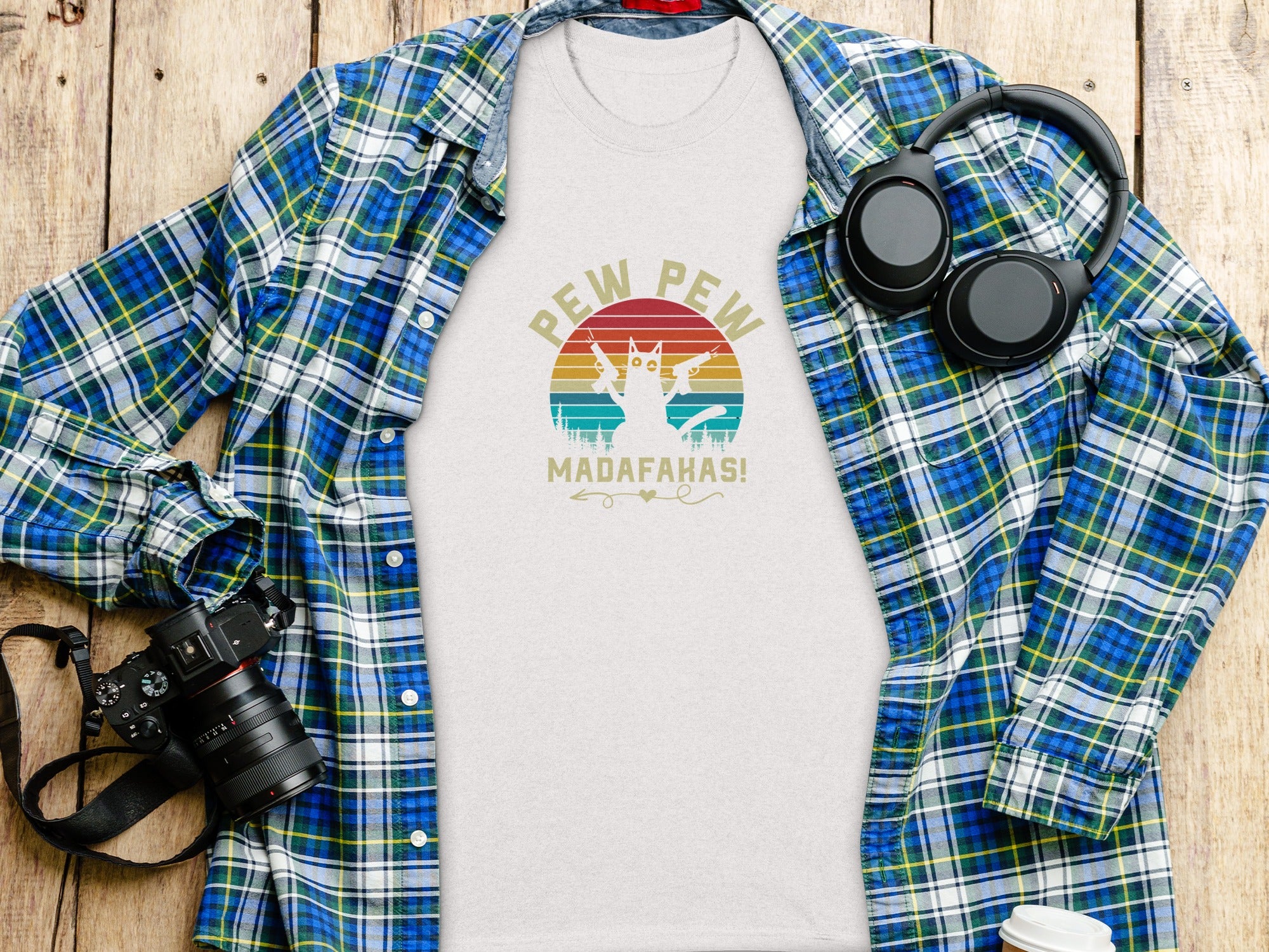 White t-shirt with cat graphic and text "PEW PEW MADAF**HAS", laid flat with a plaid shirt.
