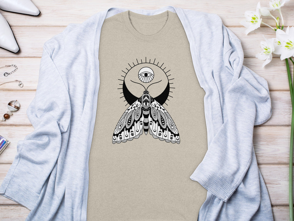 Tan t-shirt with black moth and eye graphic, draped with a light blue cardigan.