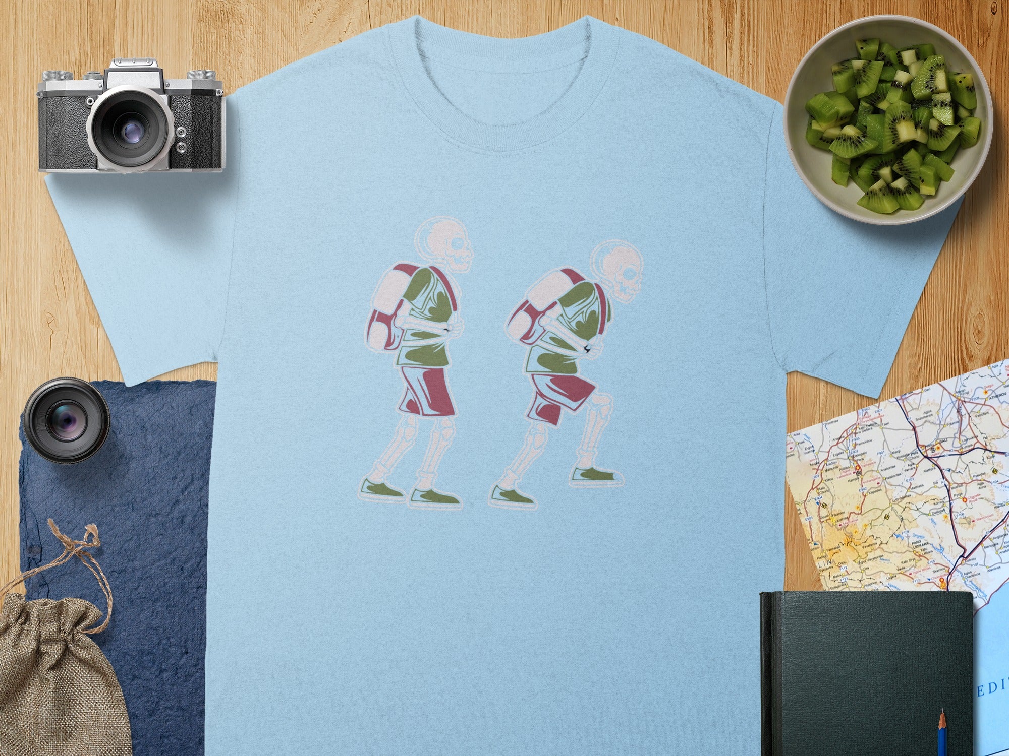Light blue t-shirt with graphic of two skeletons hiking with backpacks.