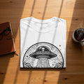 White t-shirt with black alien and UFO graphic on a wooden table.