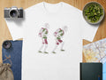 White t-shirt with two skeletons wearing backpacks and shorts, walking to the right.