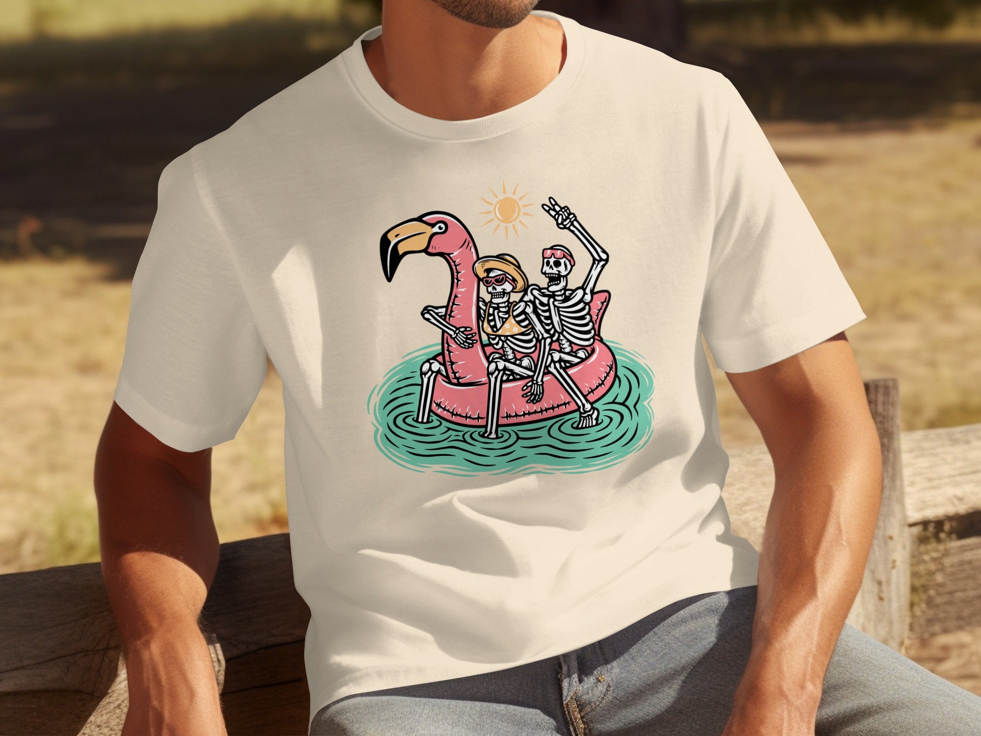 Two skeletons ride a flamingo float under a cartoon sun, on a light-colored t-shirt.