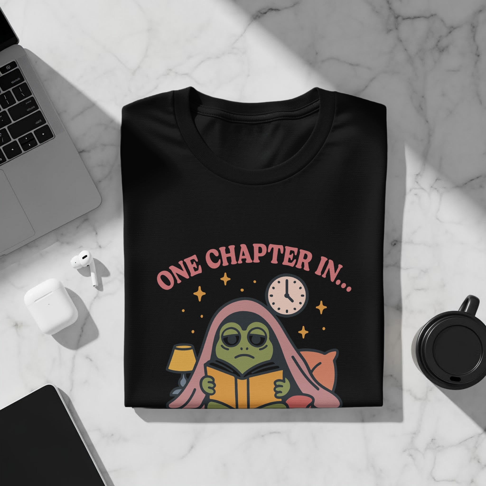 Black t-shirt with cartoon frog reading under a blanket, "One Chapter In..." text.
