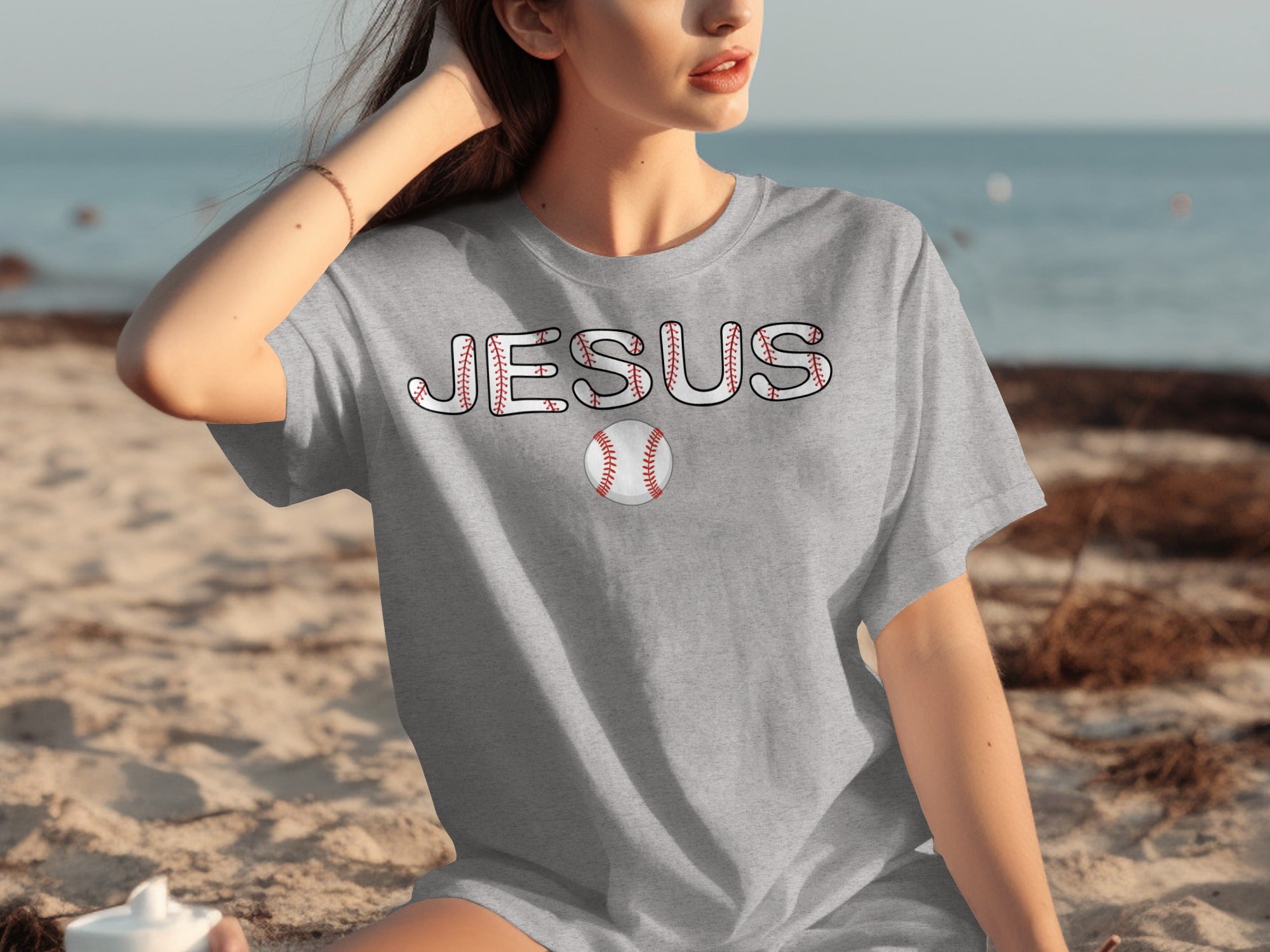 Gray t-shirt with "JESUS" text and baseball design on a beach.