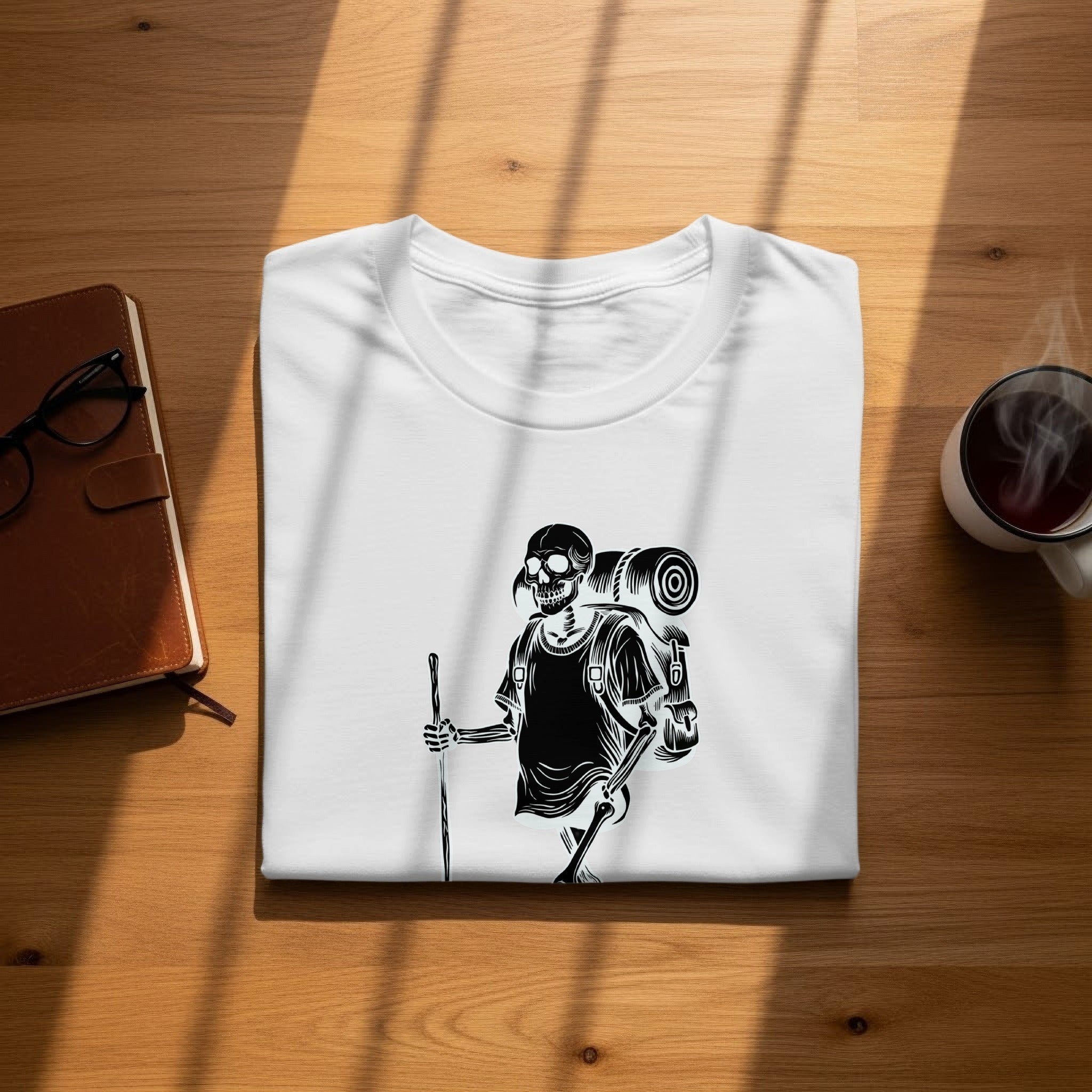 White t-shirt with black skeleton hiker graphic, laid flat on a wooden surface.