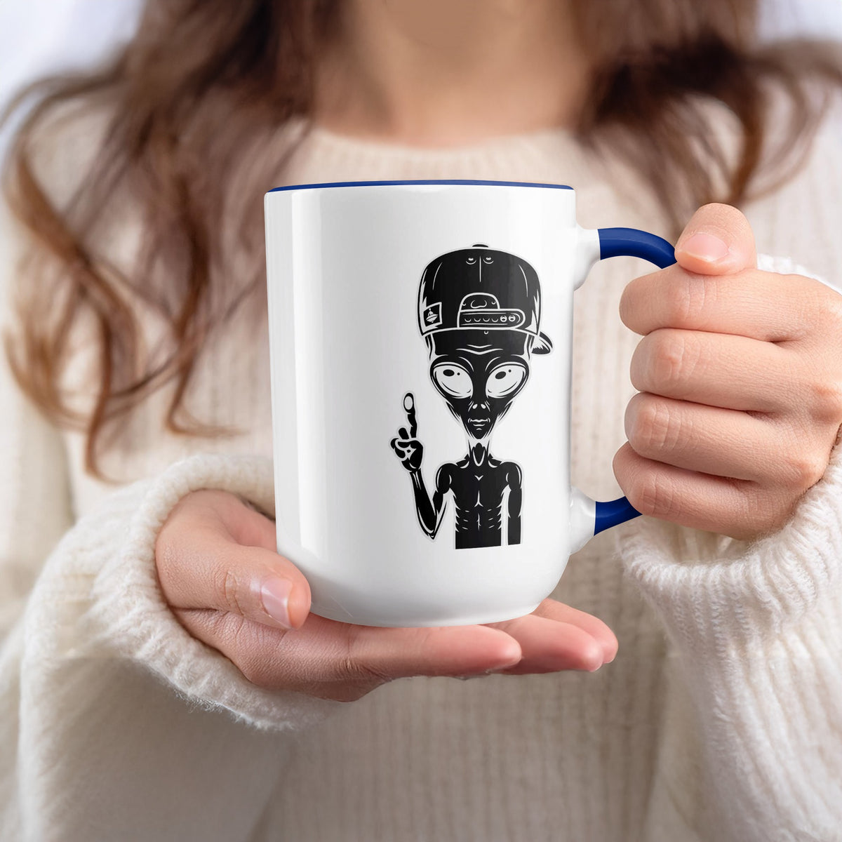 White mug with blue rim holding a black alien design wearing a baseball cap.