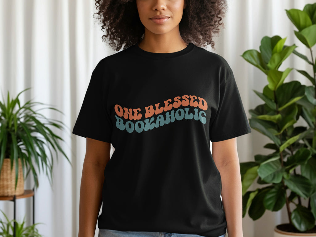 Black t-shirt with "ONE BLESSED BOOKAHOLIC" text in wavy orange and blue retro lettering.