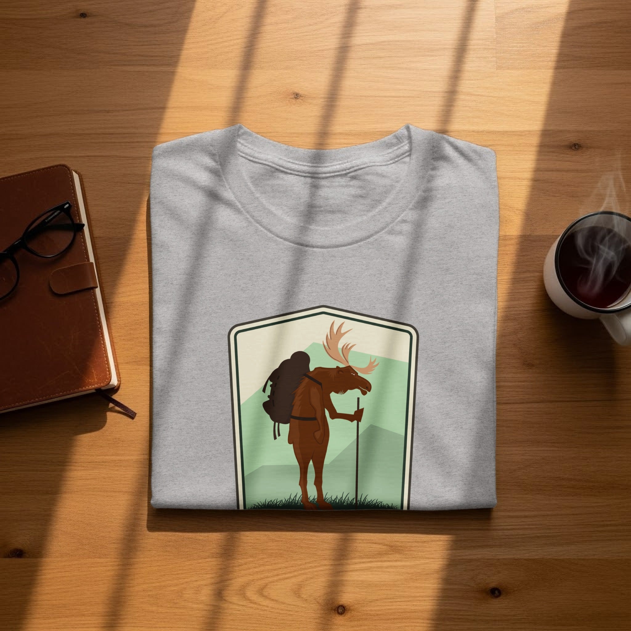Grey t-shirt with a shield logo featuring a moose hiking with a backpack and walking stick.