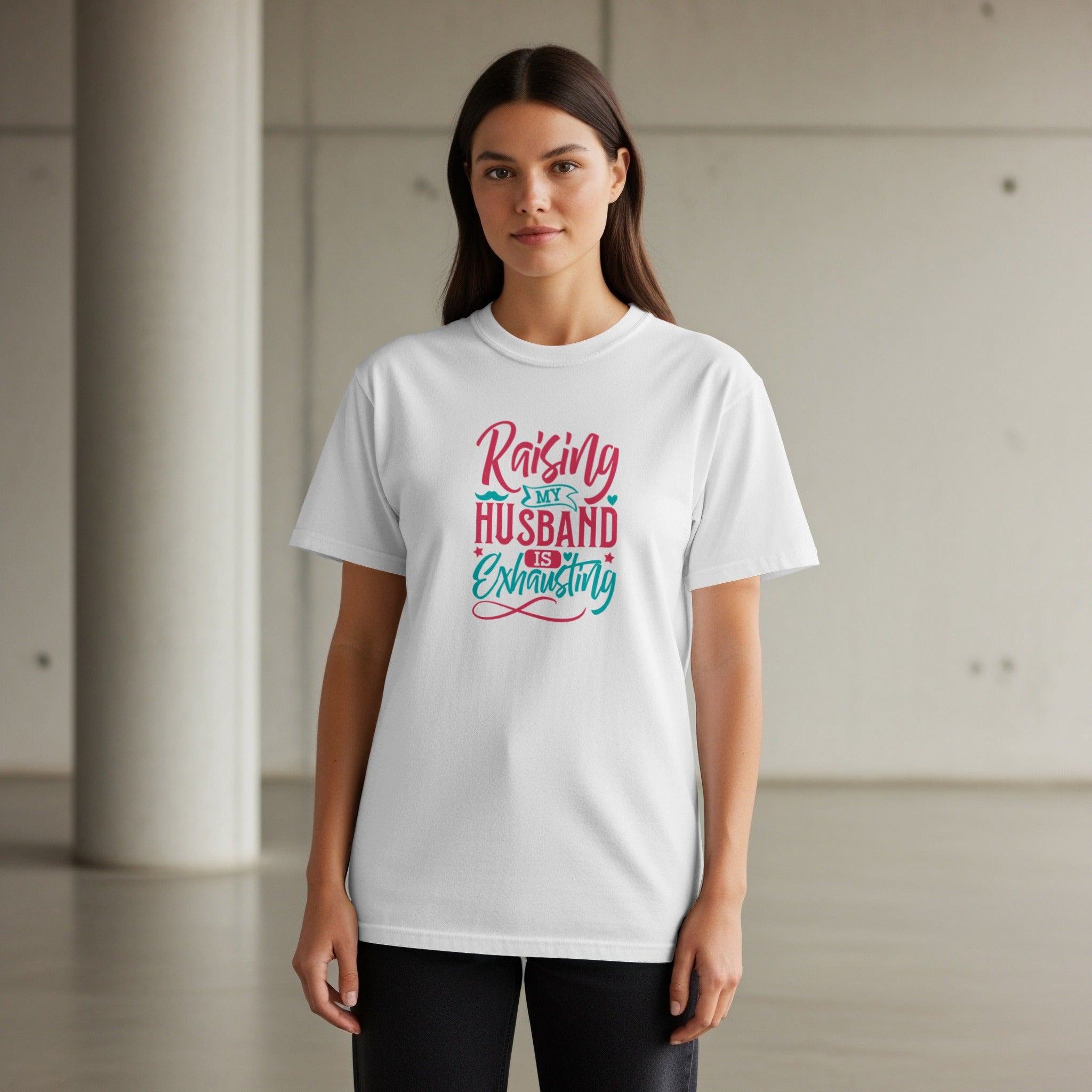 White t-shirt with pink and teal text that reads "Raising My Husband Is Exhausting".