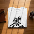 Black linocut design of a cat with a backpack hiking up a mountain on a white t-shirt.