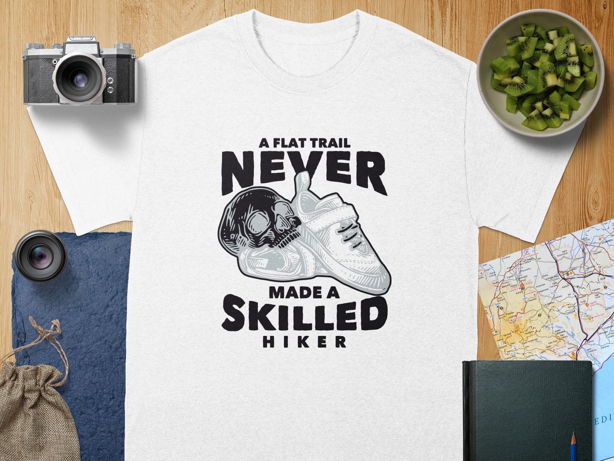 White t-shirt with skull, sneaker, and text "A flat trail never made a skilled hiker".