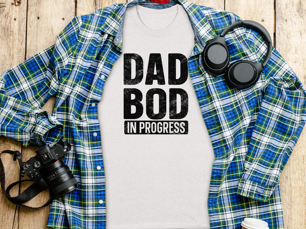 White t-shirt with "DAD BOD IN PROGRESS" text, laid flat with plaid shirt and headphones.