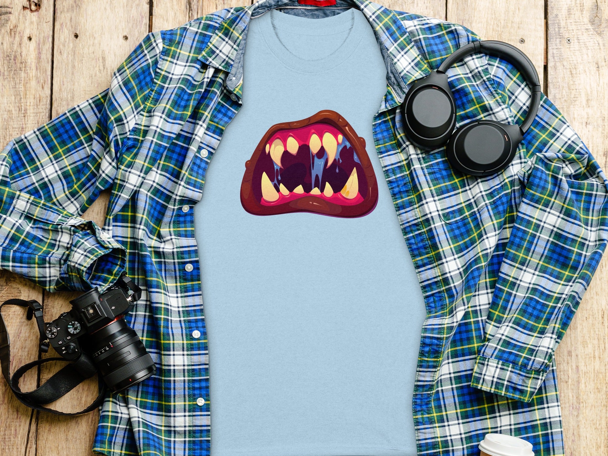 Light blue t-shirt with cartoon monster mouth graphic, laid out with flannel shirt and headphones on wood.