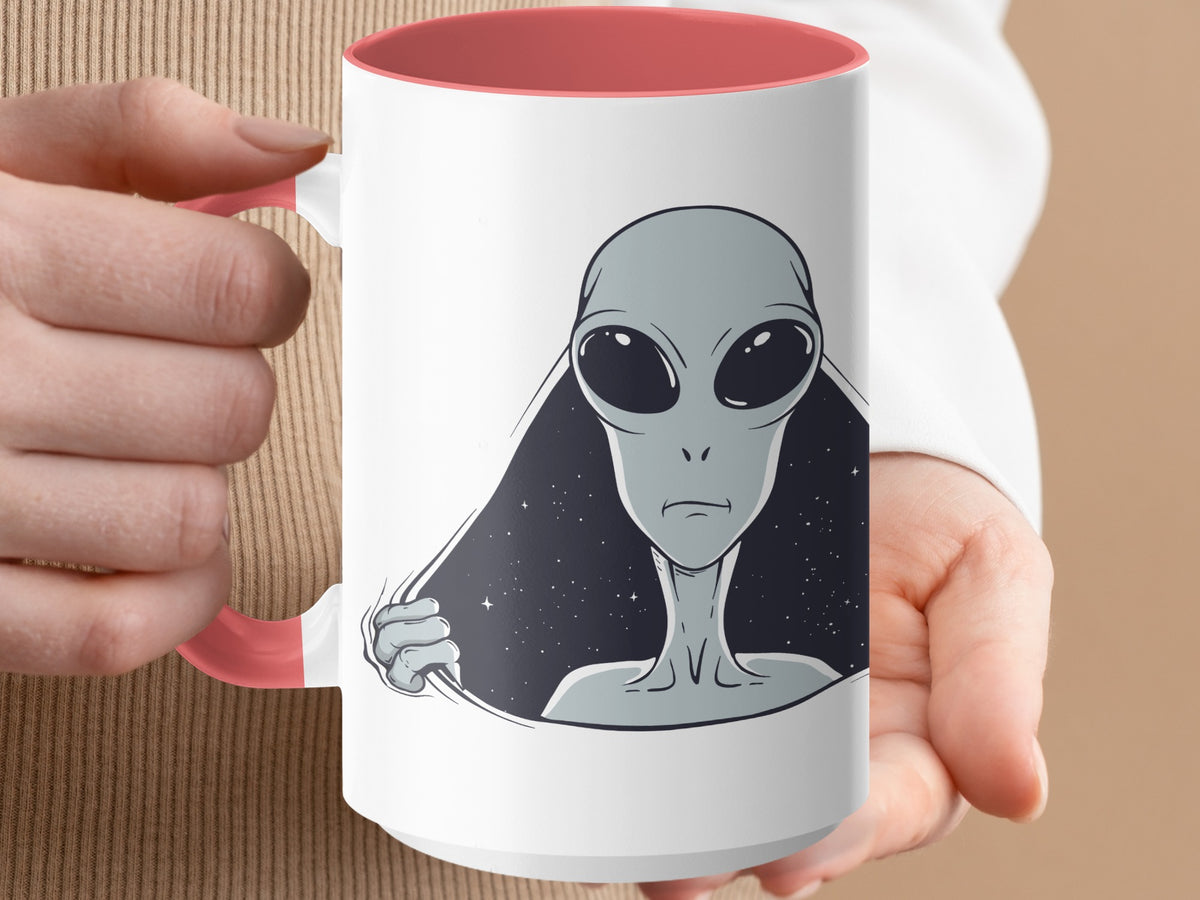 White mug with a pink handle featuring a graphic of a grey alien peeking through a starry background.