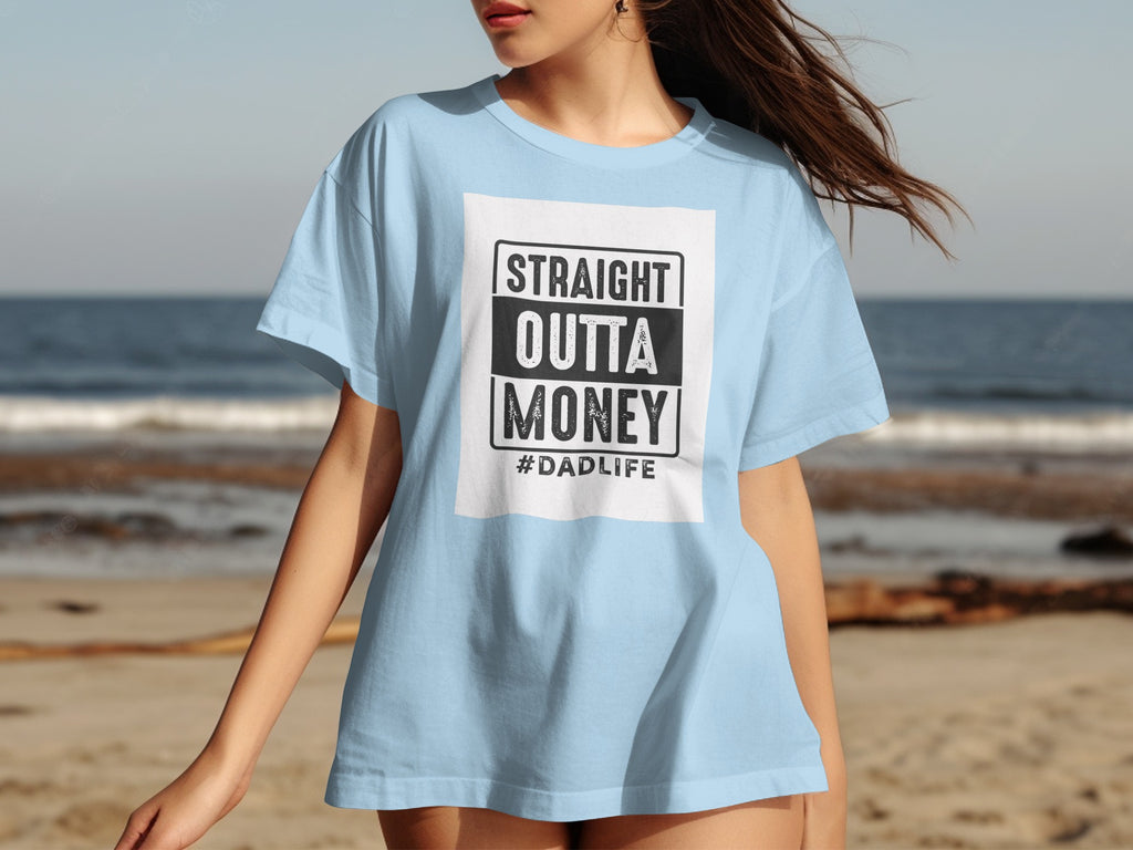 Light blue t-shirt with "STRAIGHT OUTTA MONEY #DADLIFE" graphic on a beach.