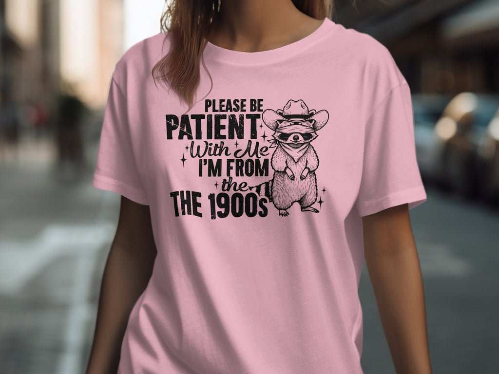 Pink t-shirt with black graphic of raccoon in cowboy hat and text "PLEASE BE PATIENT With Me I'M FROM the 1900s"
