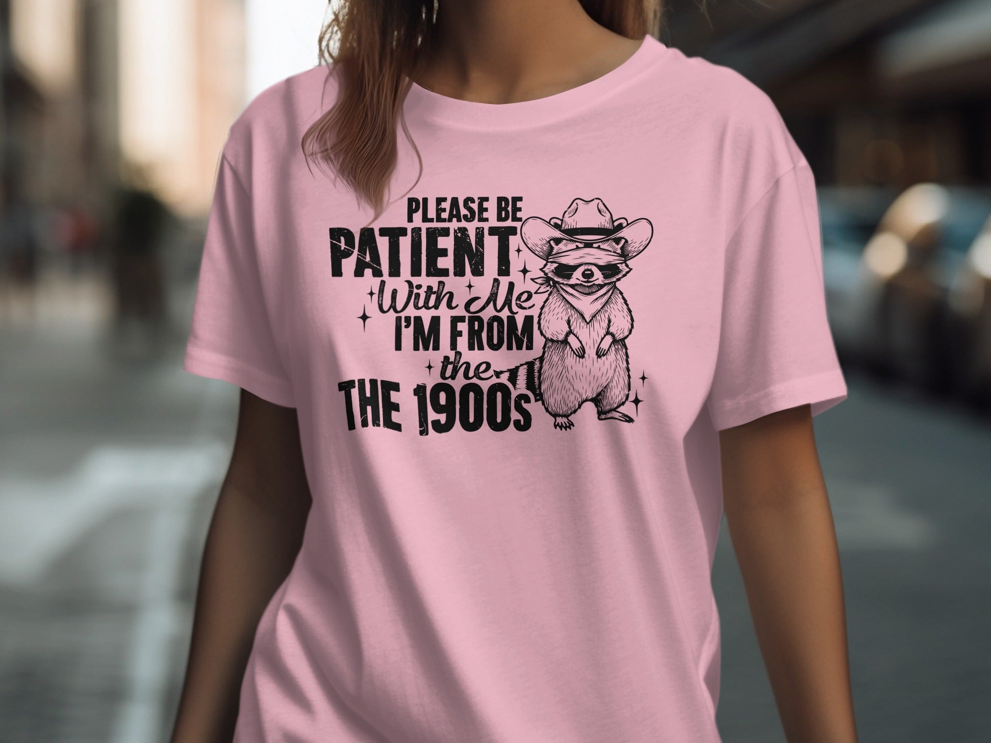 Pink t-shirt with black graphic of raccoon in cowboy hat and text "PLEASE BE PATIENT With Me I'M FROM the 1900s"