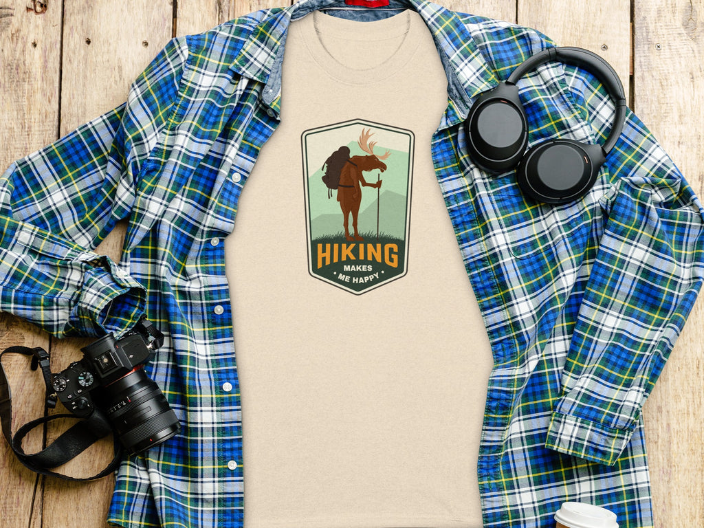 Light tan t-shirt with "Hiking Makes Me Happy" logo featuring a moose, next to plaid shirt, headphones, and camera.