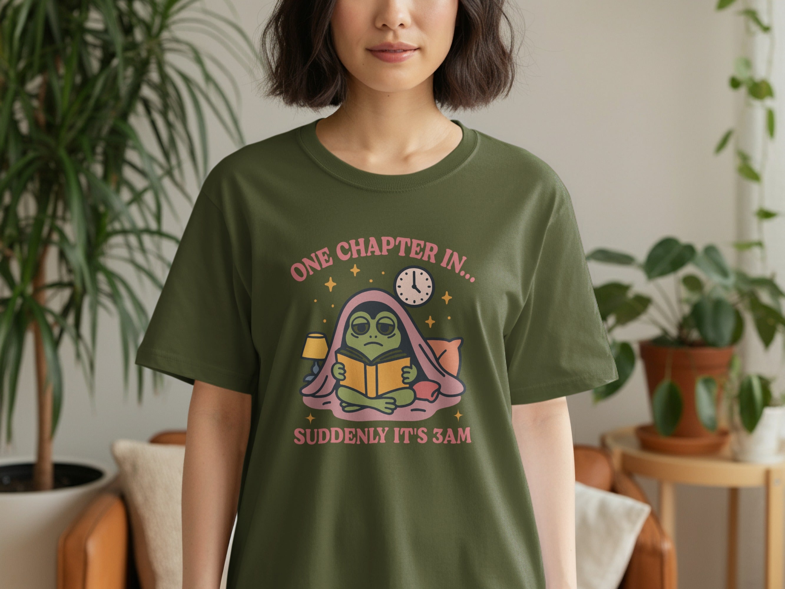 Green t-shirt with a frog reading a book under blankets, text "ONE CHAPTER IN... SUDDENLY IT'S 3AM".