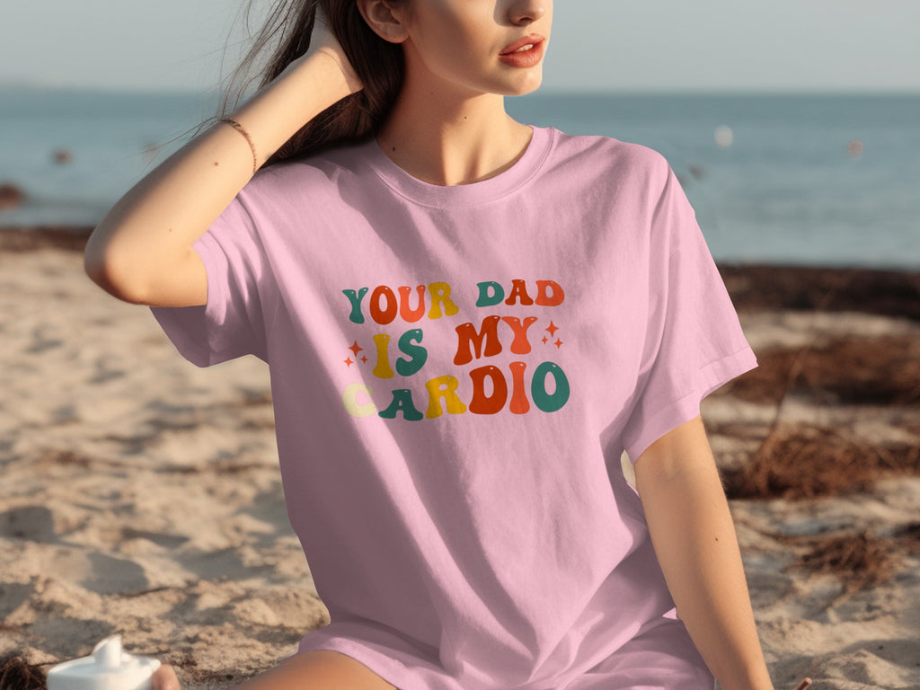 Pink t-shirt with colorful text "YOUR DAD IS MY CARDIO" on a beach.