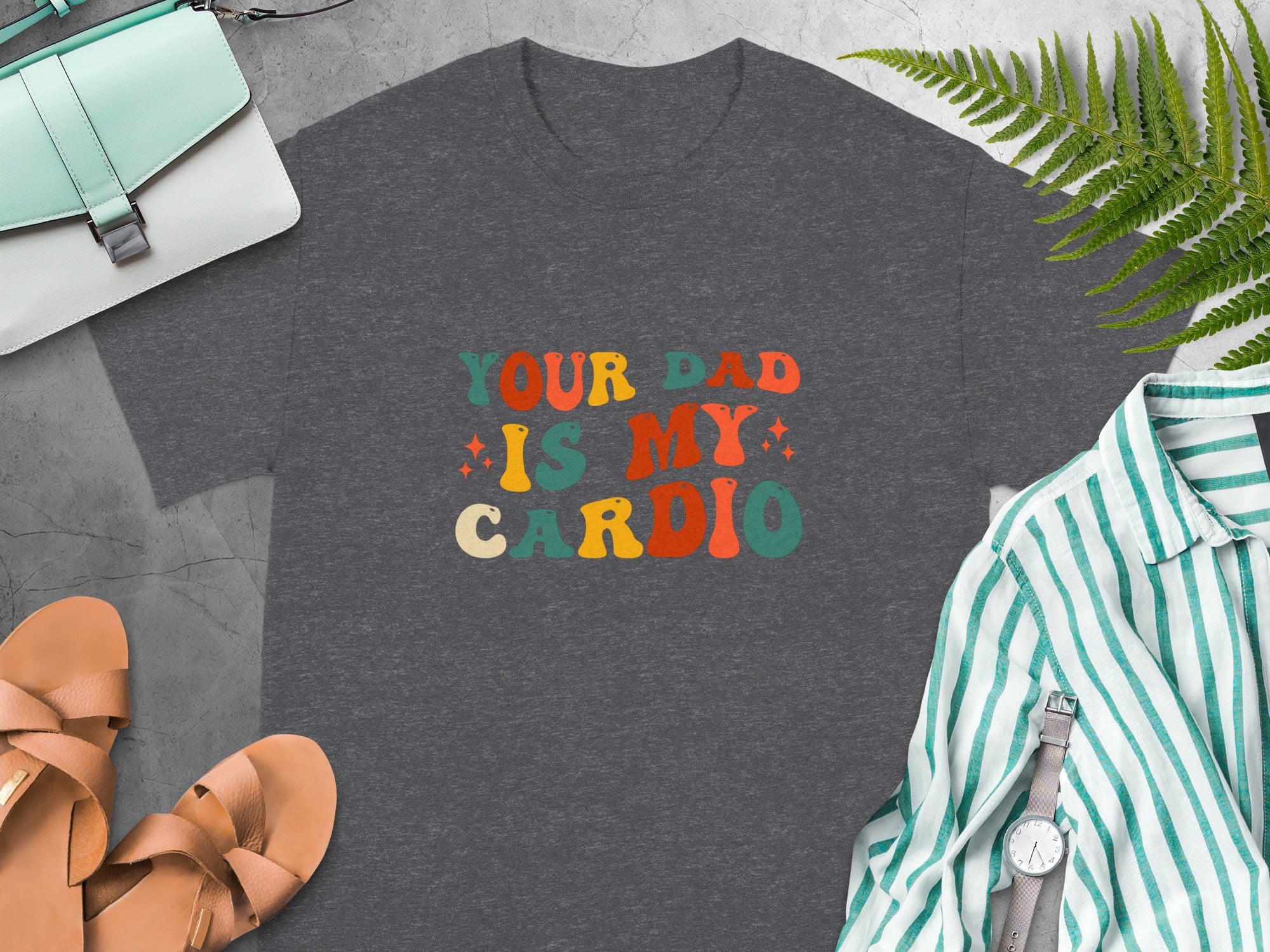 Dark gray t-shirt with colorful retro text reading "YOUR DAD IS MY CARDIO".