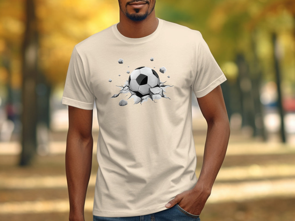 A soccer ball bursts through cracked ground on a light beige t-shirt.