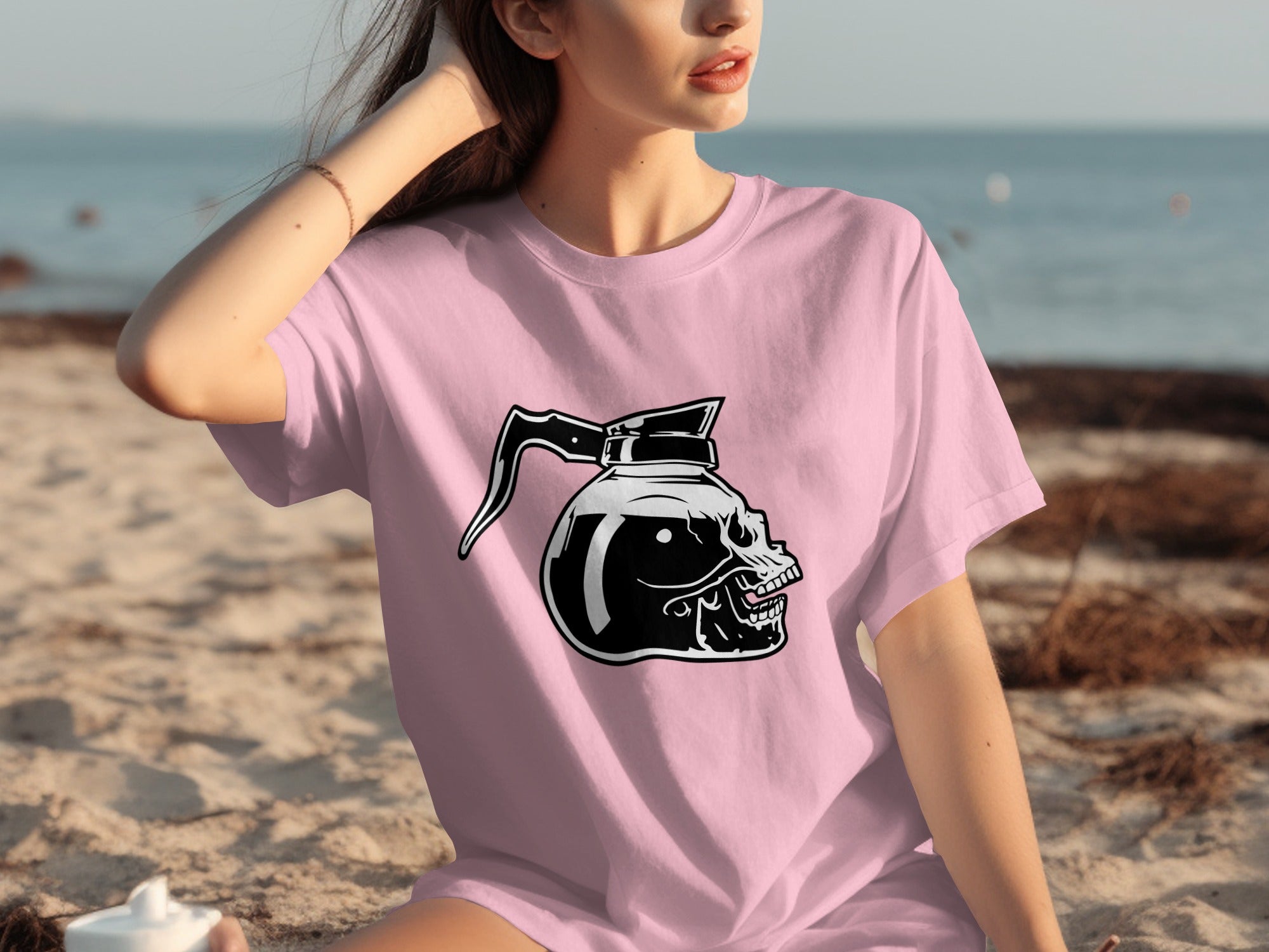 Pink t-shirt with a black graphic of a skull-shaped coffee pot on the beach.