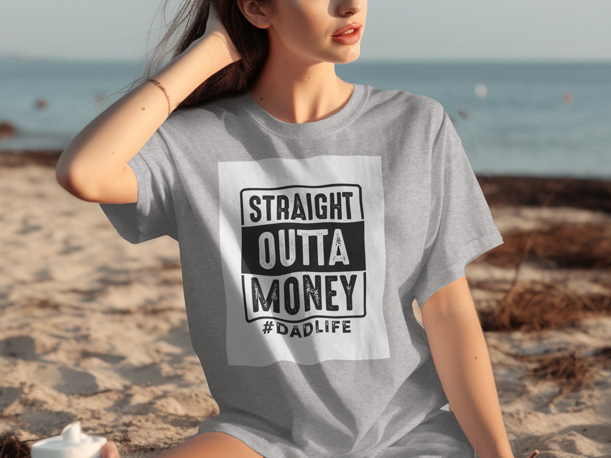 Gray t-shirt with "STRAIGHT OUTTA MONEY #DADLIFE" graphic on the beach.