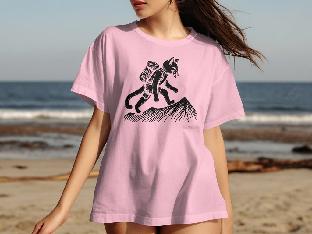 Pink t-shirt with black graphic of a cat hiking up a mountain with a backpack.