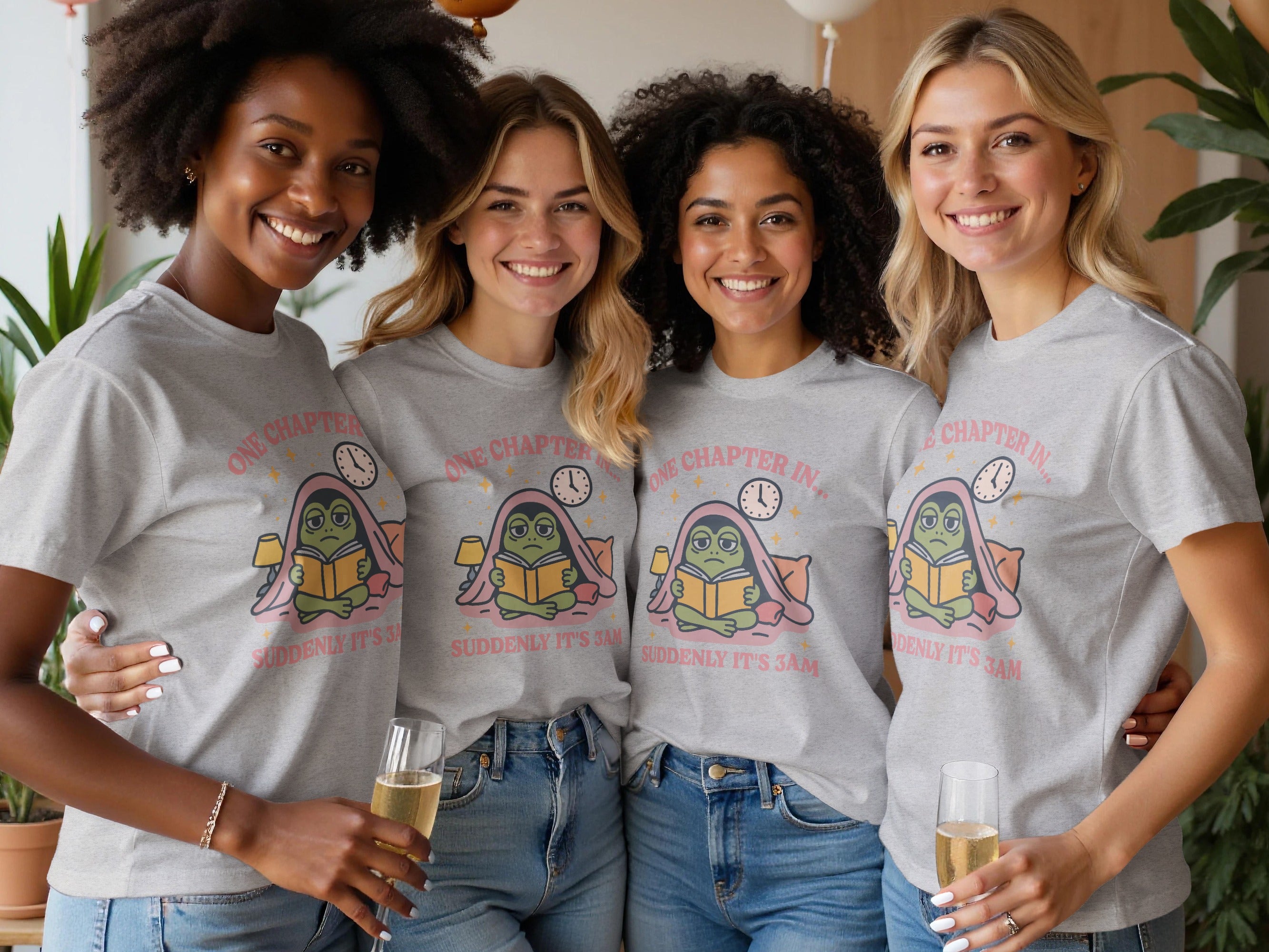 Four smiling women wear grey t-shirts with a frog reading a book design.