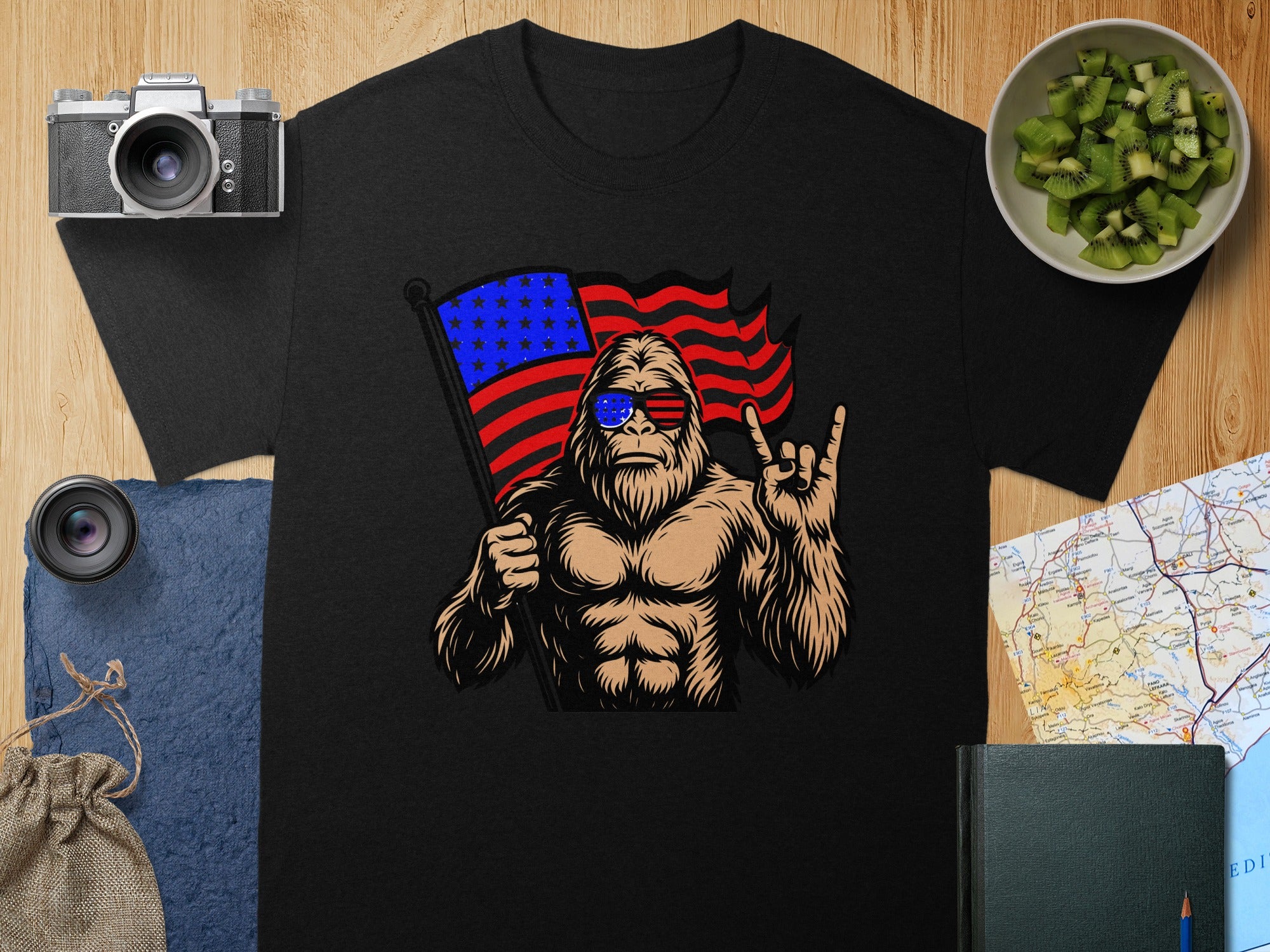 Bigfoot wearing American flag sunglasses holds flag, raises rock hand gesture.