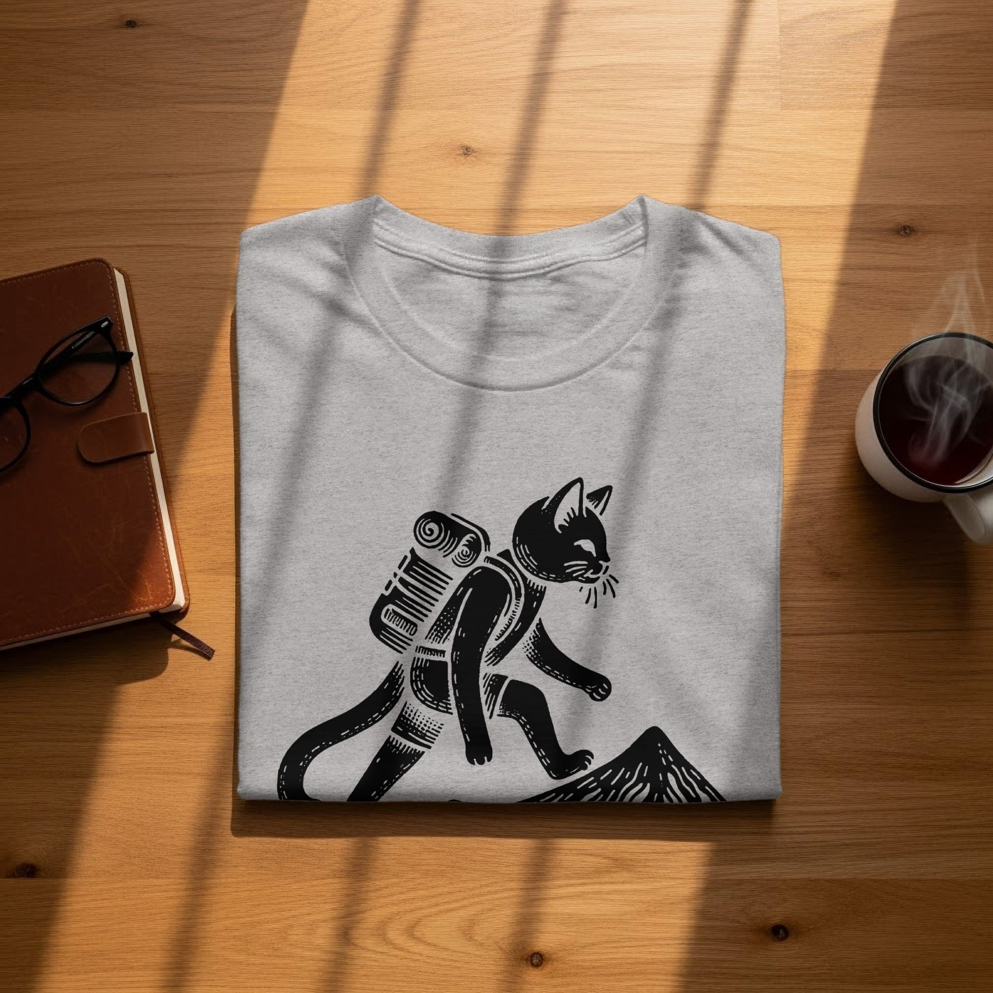 Light grey t-shirt with black linocut design of a cat with a backpack hiking up a mountain.