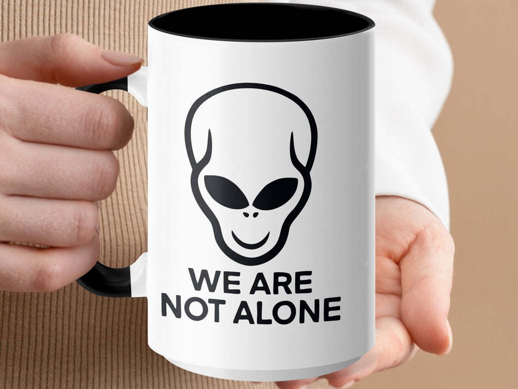 White mug with black rim, handle, alien graphic, and "WE ARE NOT ALONE" text.